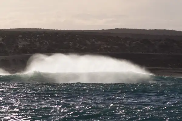 Vue du large de la houle venant se briser sur un rivage en gros rouleaux. Le vent lève des embruns à revers de la vague.