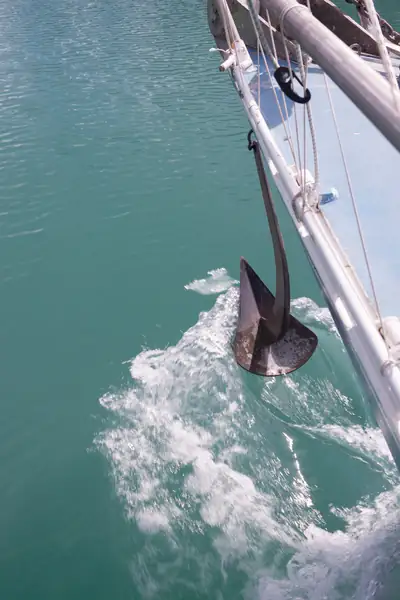 Vue de l'étrave d'un voilier, prise du bord, alors qu'il avance à bonne vitesse. De sa proue pend l'ancre du bateau, qui vient glisser à la surface de l'eau.