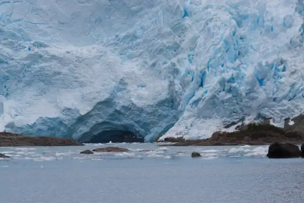 Glacier vêlant dans un bras de mer. Devant lui, des roches émergent à peine de l'eau et des morceaux de glace flottante sont échoués sur les hauts-fonds.