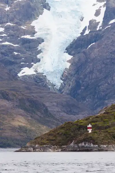Une langue glacière descend de la montage en s'accrochant aux parois rocheuses. En avant-plan, le long d'un fjord glaciaire de la Patagonie chilienne, un petit feu blanc et rouge permet d'aider la navigation dans les canaux.