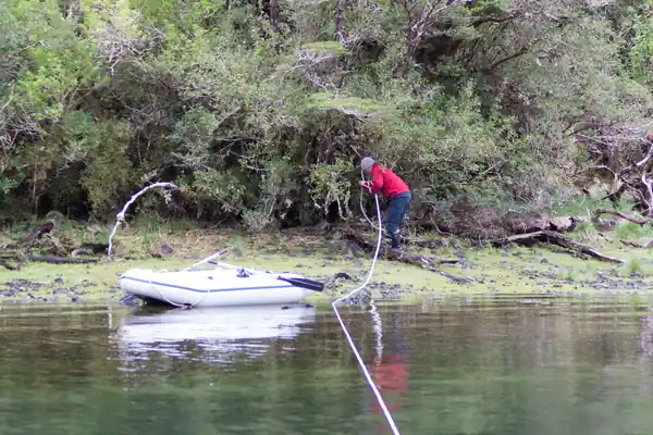 Un équipier va porter à terre une aussière flottante. Il a débarqué sur une petite plage étroite en pente douce et couverte de végétaux. L'annexe, avirons encore à poste, est simplement posée sur le rivage.