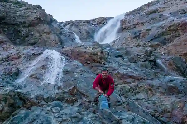 Une cascade d'eau glaciale dévale une pente rocailleuse. Une personne y remplit un bidon d'eau.