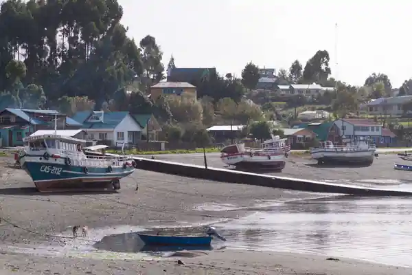 Rivage de l'île de Chiloé à marée basse. Une cale s'avance en descendant vers l'eau tandis que des bateaux de pêche sont échoués devant le rivage et les maisons d'un village.